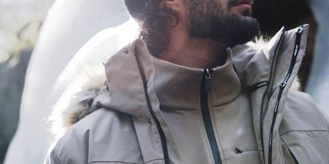 Uomo con giacca marrone, pantaloni neri e berretto grigio cammina attraverso una fitta foresta. Indossa guanti neri e uno zaino. La foresta ha alberi alti e foglie cadute. Logo "Rukka" in alto.