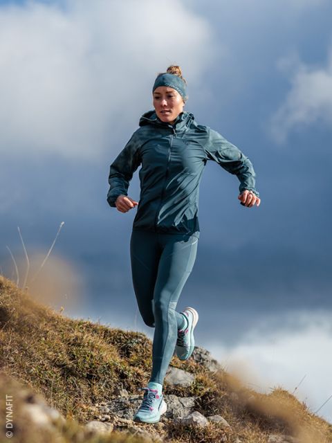 Una donna in abbigliamento sportivo color turchese corre in discesa su un sentiero roccioso, con un cielo nuvoloso sullo sfondo.