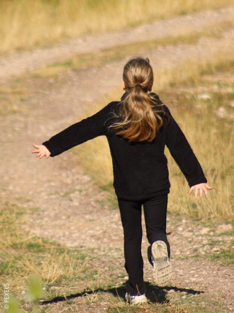 Una ragazza con i capelli lunghi e un vestito nero corre felicemente con le braccia aperte lungo un sentiero sterrato fiancheggiato dall'erba.