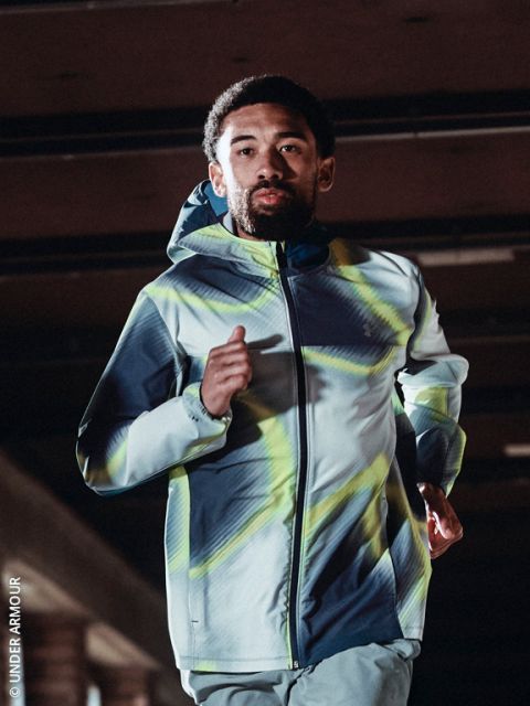Una persona con una felpa con cappuccio e scarpe da corsa fa jogging lungo un sentiero innevato in una giornata fredda e limpida, con lo skyline della città sullo sfondo.