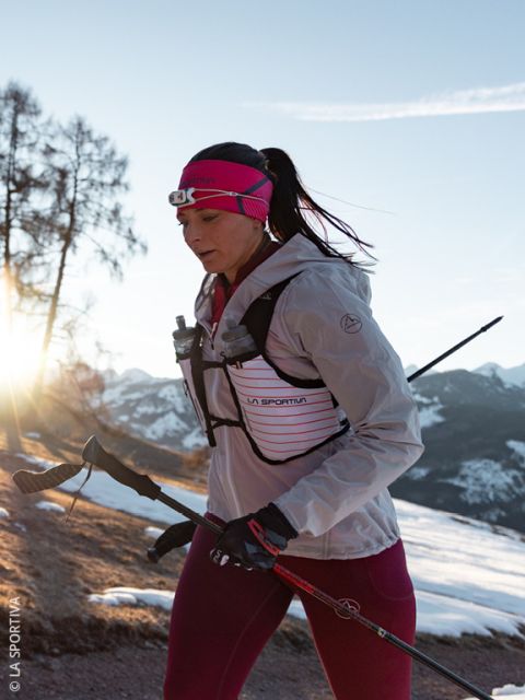 Una donna che indossa una giacca e guanti rosa-grigi corre in un paesaggio montano innevato e porta due bottiglie d'acqua nelle tasche anteriori del suo gilet da corsa.