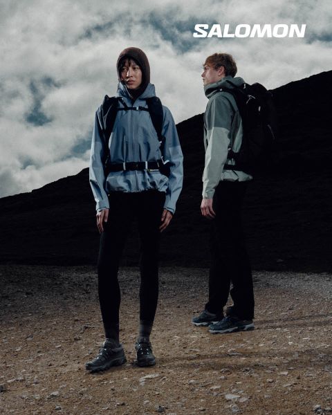 Primo piano di una persona che indossa scarpe Salomon e calze Salomon con gradiente blu. Si trova su un prato con un pittoresco paesaggio montano sullo sfondo. Un escursionista in abbigliamento outdoor si trova più lontano, anch'esso sul prato.