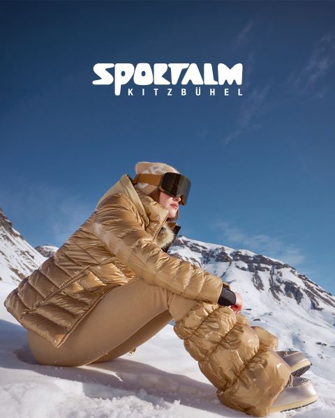Una persona con un piumino metallizzato e pantaloni neri è sdraiata su un terreno innevato con montagne sullo sfondo sotto un cielo nuvoloso. La scena è tranquilla e invernale. Nell'angolo in alto a destra si legge il testo "SPORTALM KITZBÜHEL".