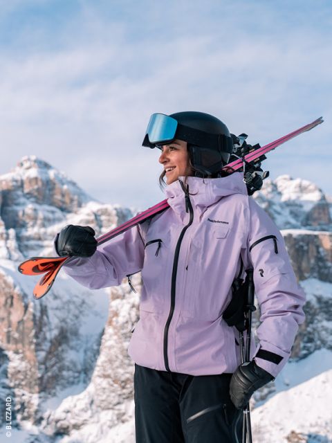 Una sciatrice con una giacca color lavanda e un casco sorride mentre porta gli sci sulla spalla; sullo sfondo si vedono montagne innevate.