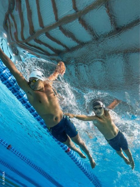 Due nuotatori con occhialini e cuffie da nuoto gareggiano sott'acqua in una piscina; uno allunga la mano verso l'altro vicino alla corsia di separazione.