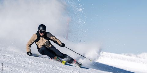 Uno sciatore con una giacca beige e un casco nero fa curve strette su un pendio innevato, sollevando una nuvola di neve contro il cielo azzurro brillante.