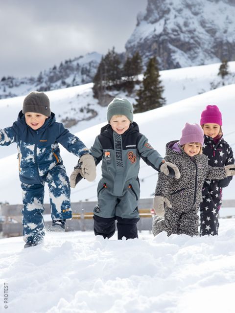 Quattro bambini in abiti invernali giocano e sorridono nella neve, con montagne e alberi innevati visibili sullo sfondo.