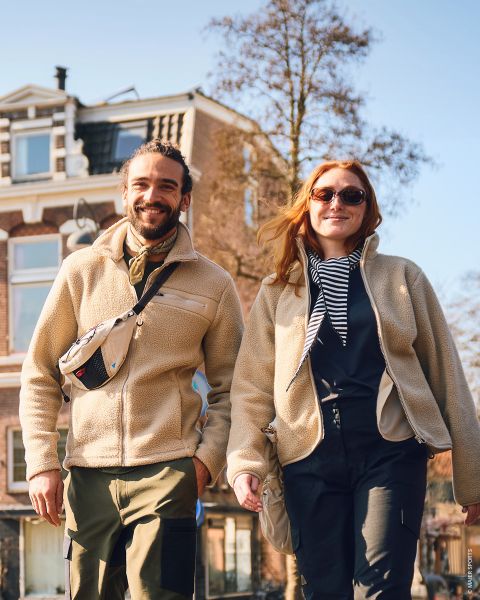 Un uomo e una donna sorridenti camminano all'aperto in una giornata di sole, vestiti con giacche e stivali casual, tenendosi per mano, con un grande edificio in mattoni sullo sfondo.