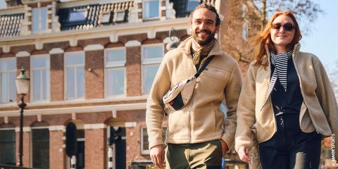 Un uomo e una donna sorridenti camminano mano nella mano su un ponte in una giornata di sole, con edifici storici visibili sullo sfondo.