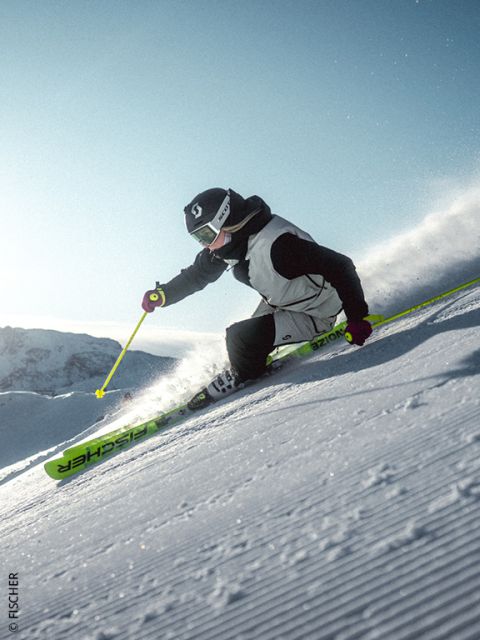 Uno sciatore in giacca blu e con casco scende un pendio montuoso innevato al tramonto, sollevando neve fresca, con cime montuose aspre e illuminate dal sole sullo sfondo.