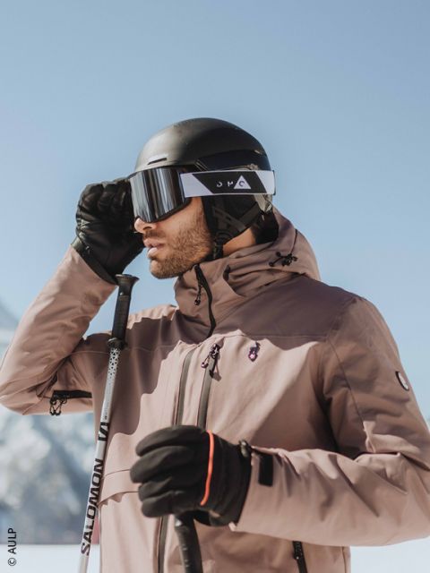 Primo piano di una persona che indossa un casco da sci grigio, occhiali da sci riflettenti e una giacca scura, con un paesaggio innevato sfocato sullo sfondo.