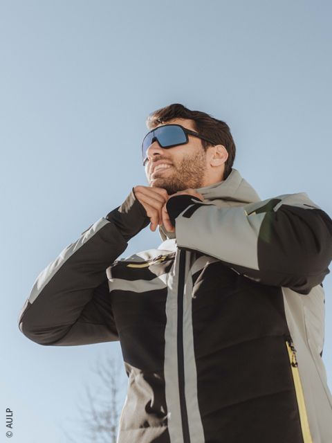 Uno sciatore che indossa un casco bianco, occhiali riflettenti, una giacca e guanti marrone-neri chiude la sua giacca all'aperto con montagne innevate sullo sfondo.