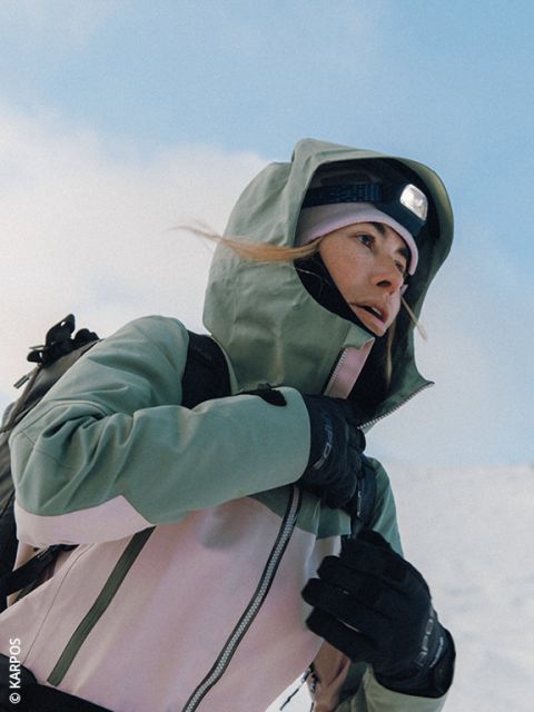 Una donna che indossa una giacca Dynafit imbottita, occhiali da sole e guanti si prepara per un'escursione in un paesaggio montano innevato, con cime innevate sullo sfondo. Porta uno zaino e bastoncini da sci.