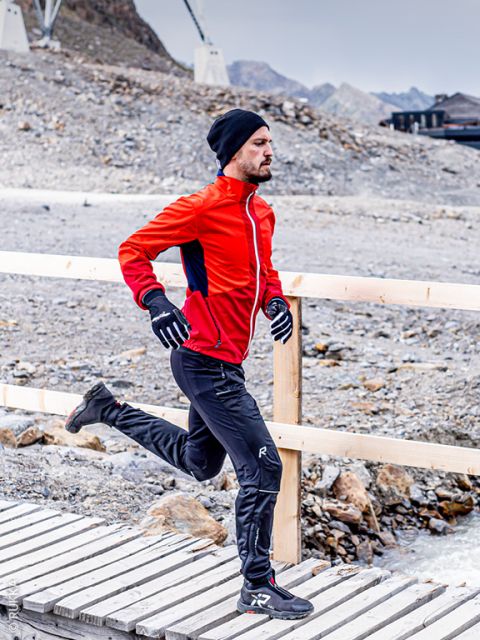 Un uomo con una giacca rossa, pantaloni neri, guanti e un cappello cammina su un ponte di legno in una zona rocciosa e montuosa.