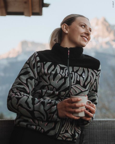 Una donna che indossa una giacca in pile fantasia con cerniera tiene in mano una tazza e guarda le montagne sullo sfondo.