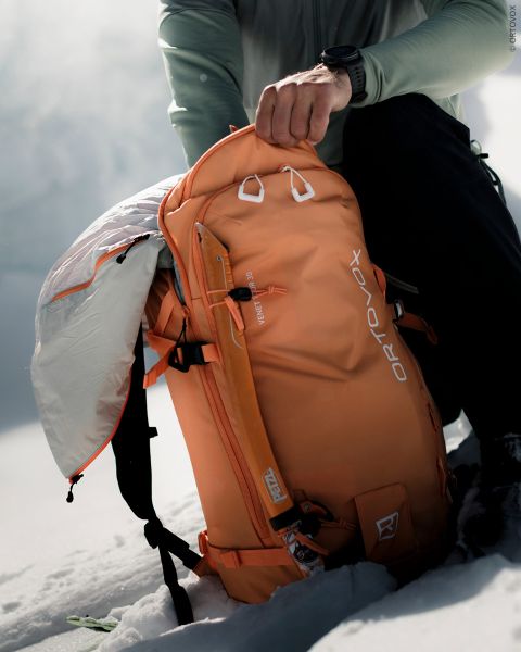 Un escursionista apre uno zaino Ortovox arancione in un paesaggio montano innevato.