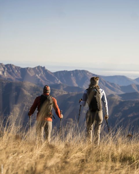 Due escursionisti con zaini e bastoni da trekking si trovano nell'erba alta e guardano catene montuose lontane sotto un cielo limpido.